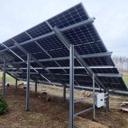 SOLMAX - Montaż paneli fotowoltaicznych na konstrukcji stalowej, widok od spodu, z widocznym inwerterem i młodą choinką w tle.