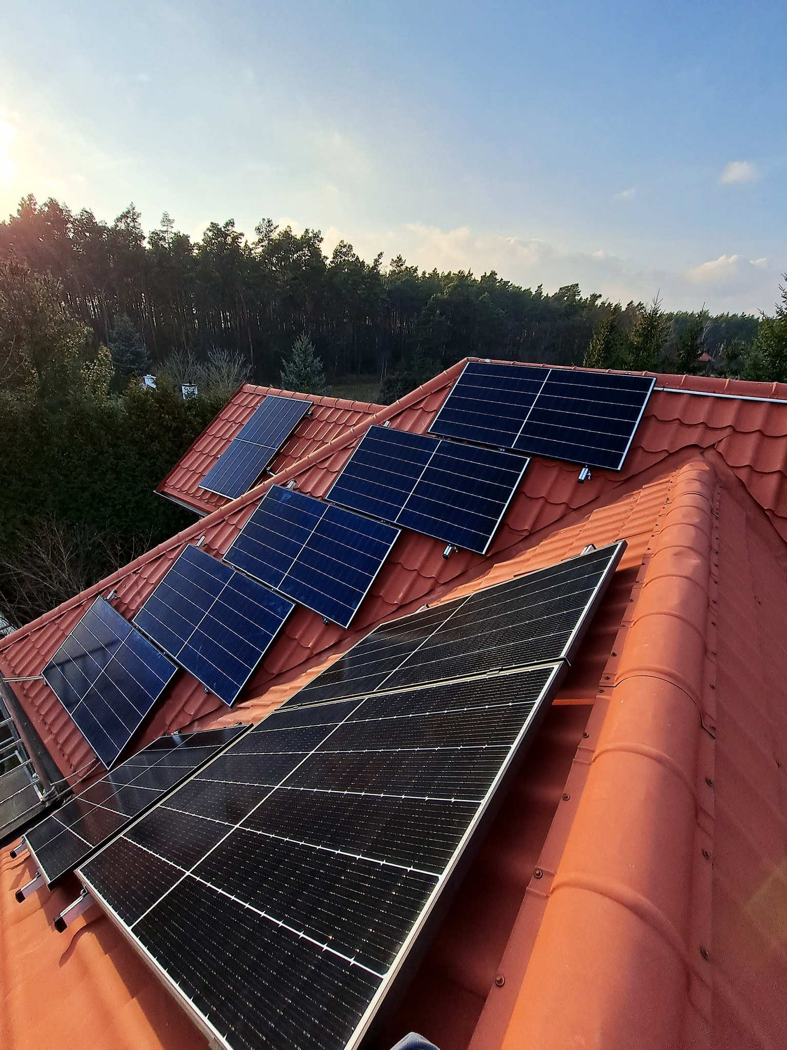 Panele słoneczne zamontowane na dachu z czerwonej dachówki, widok z góry, w tle las i błękitne niebo.