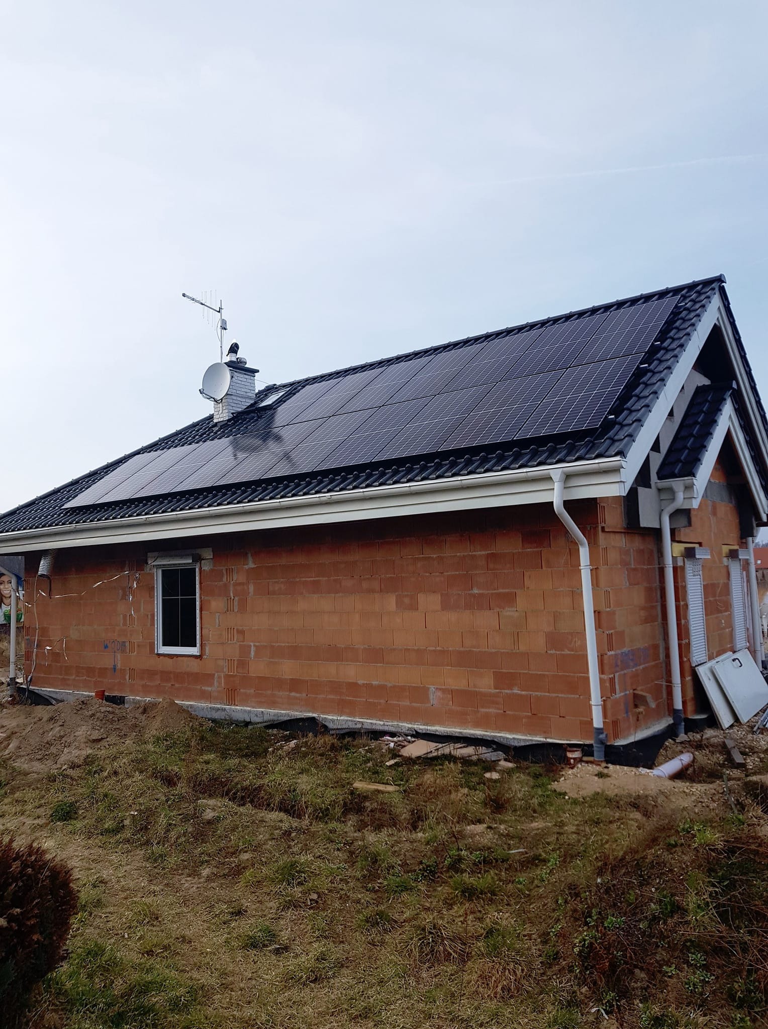 Dom w Łodzi z panelami fotowoltaicznymi na dachu, widoczna antena satelitarna i komin. Budynek w trakcie budowy, z surowymi ścianami z cegły.