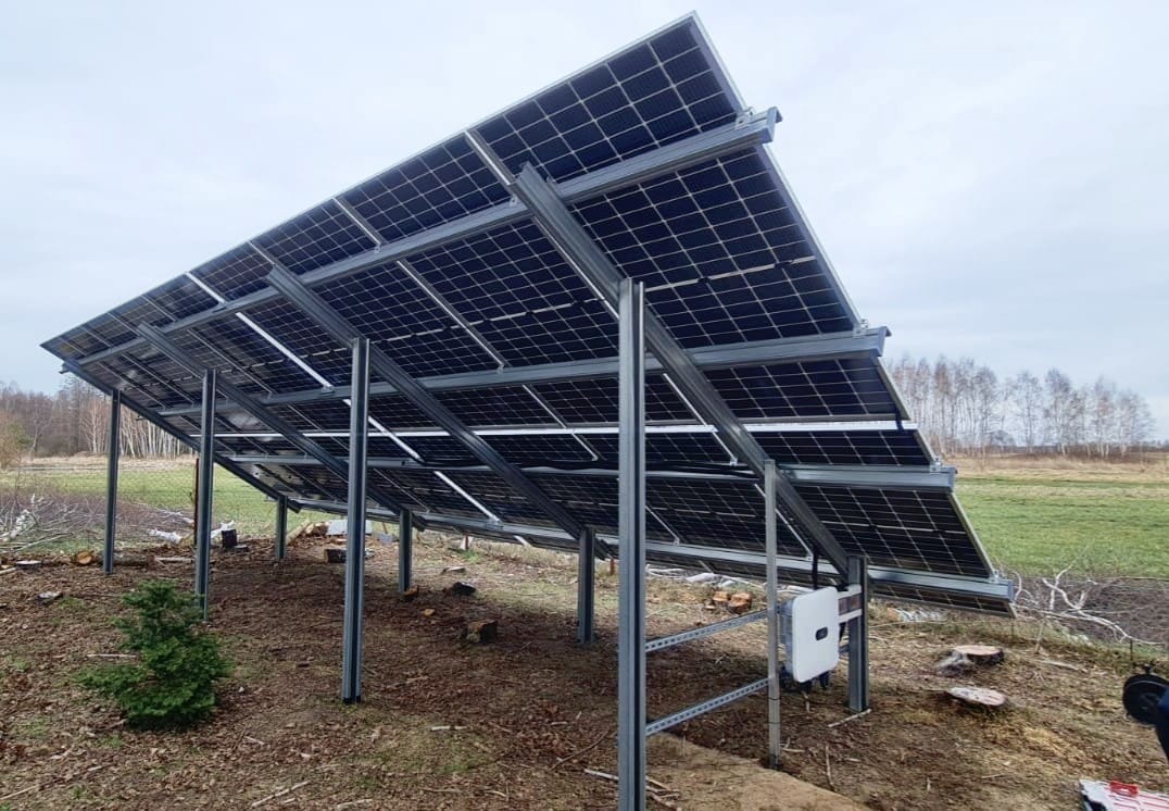 Montaż paneli fotowoltaicznych na konstrukcji stalowej, widok od spodu, z widocznym inwerterem i młodą choinką w tle.