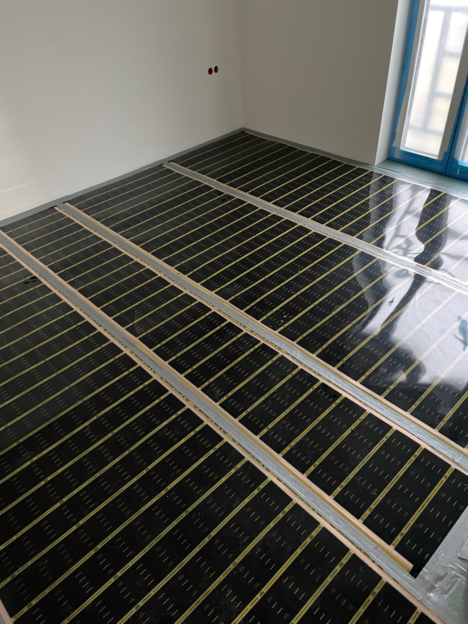 Rozłożona mata grzewcza na podłodze w nowym budownictwie, widoczne czarne panele z żółtymi paskami i metalowe listwy mocujące, okno zabezpieczone folią malarską.