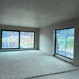 Maniak-Piotr Maniak - Surowe wnętrze w trakcie prac wykończeniowych: świeżo otynkowane ściany i sufit, z oknami zabezpieczonymi folią ochronną, widoczne wystające przewody elektryczne i listwa przypodłogowa. Podłoże...