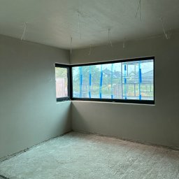 Maniak-Piotr Maniak - Pokój po tynkowaniu maszynowym ścian i sufitu, z dużym oknem zabezpieczonym folią i taśmą malarską, widoczne kable wystające z sufitu, podłoga w trakcie prac.