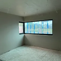 Pokój po tynkowaniu maszynowym ścian i sufitu, z dużym oknem zabezpieczonym folią i taśmą malarską, widoczne kable wystające z sufitu, podłoga w trakcie prac.