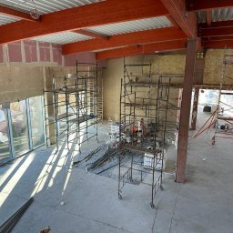 Maniak-Piotr Maniak - Wnętrze w trakcie remontu: widok z góry na liczne rusztowania, przygotowane ściany pod tynk i duże okna z widokiem na zieleń.