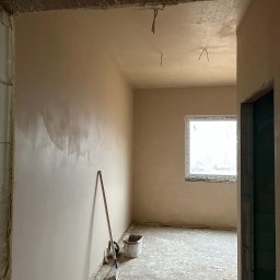 Maniak-Piotr Maniak - Wnętrze w trakcie prac wykończeniowych: ściany pokryte świeżo nałożoną, jasną warstwą tynku, widoczne narzędzia (szpachelka, wiadro) oparte o ścianę, surowy betonowy strop i podłoga, okno w tle.