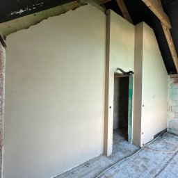 Maniak-Piotr Maniak - Świeżo otynkowane ściany wewnątrz poddasza, widoczne drewniane elementy konstrukcyjne dachu i instalacja elektryczna na podłodze.