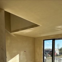 Maniak-Piotr Maniak - Wnętrze pomieszczenia po nałożeniu tynku, widoczne gładkie ściany w odcieniach beżu, nietypowe wnęki sufitowe, okno z widokiem na plac budowy i zaparkowane samochody.