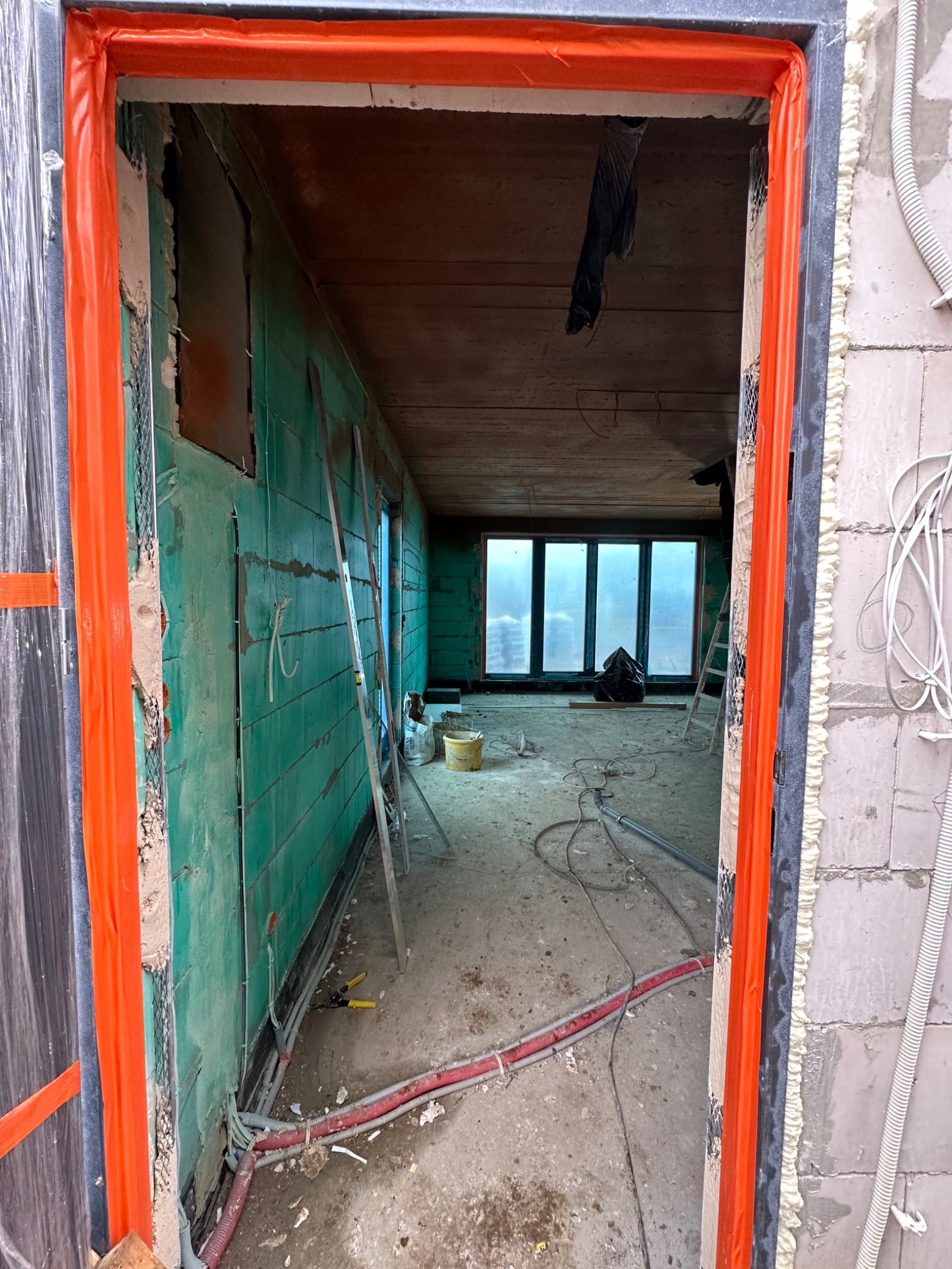 Wnętrze w trakcie remontu z widoczną ścianą obłożoną płytami gipsowo-kartonowymi, zabezpieczone pomarańczową folią ościeżnice, instalacje elektryczne na podłodze i okno z widokiem na zewnątrz.