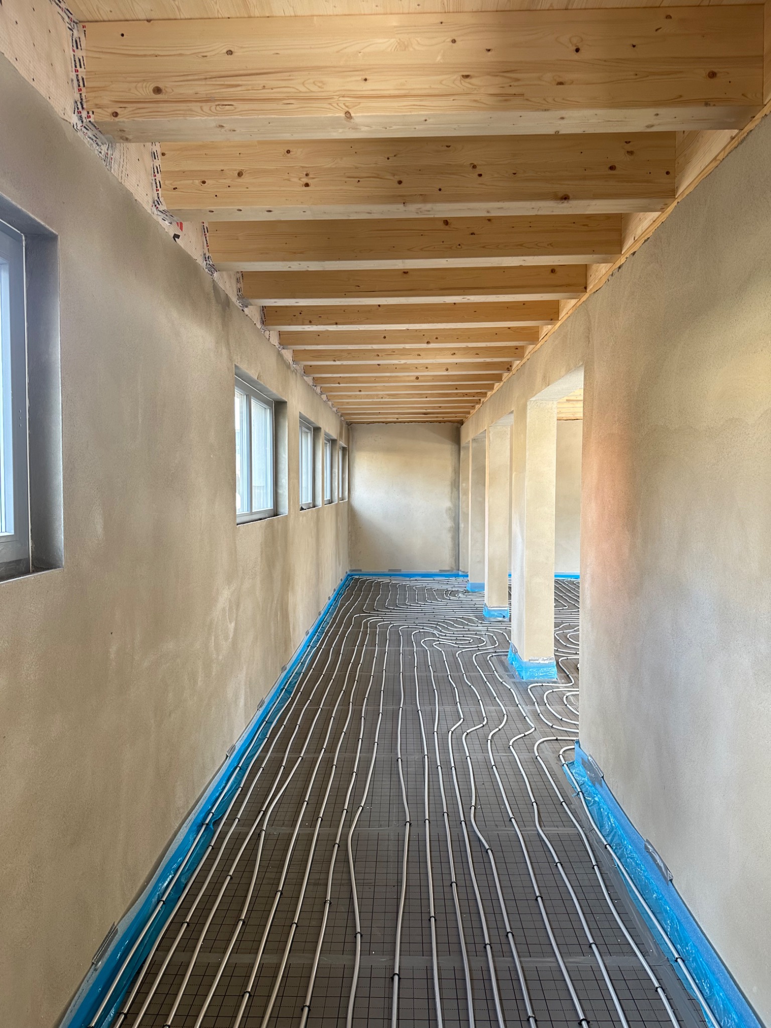 Wnętrze w trakcie prac wykończeniowych: ściany po nałożeniu tynku, widoczne drewniane belki stropowe i instalacja ogrzewania podłogowego na podłodze, zabezpieczona folią.