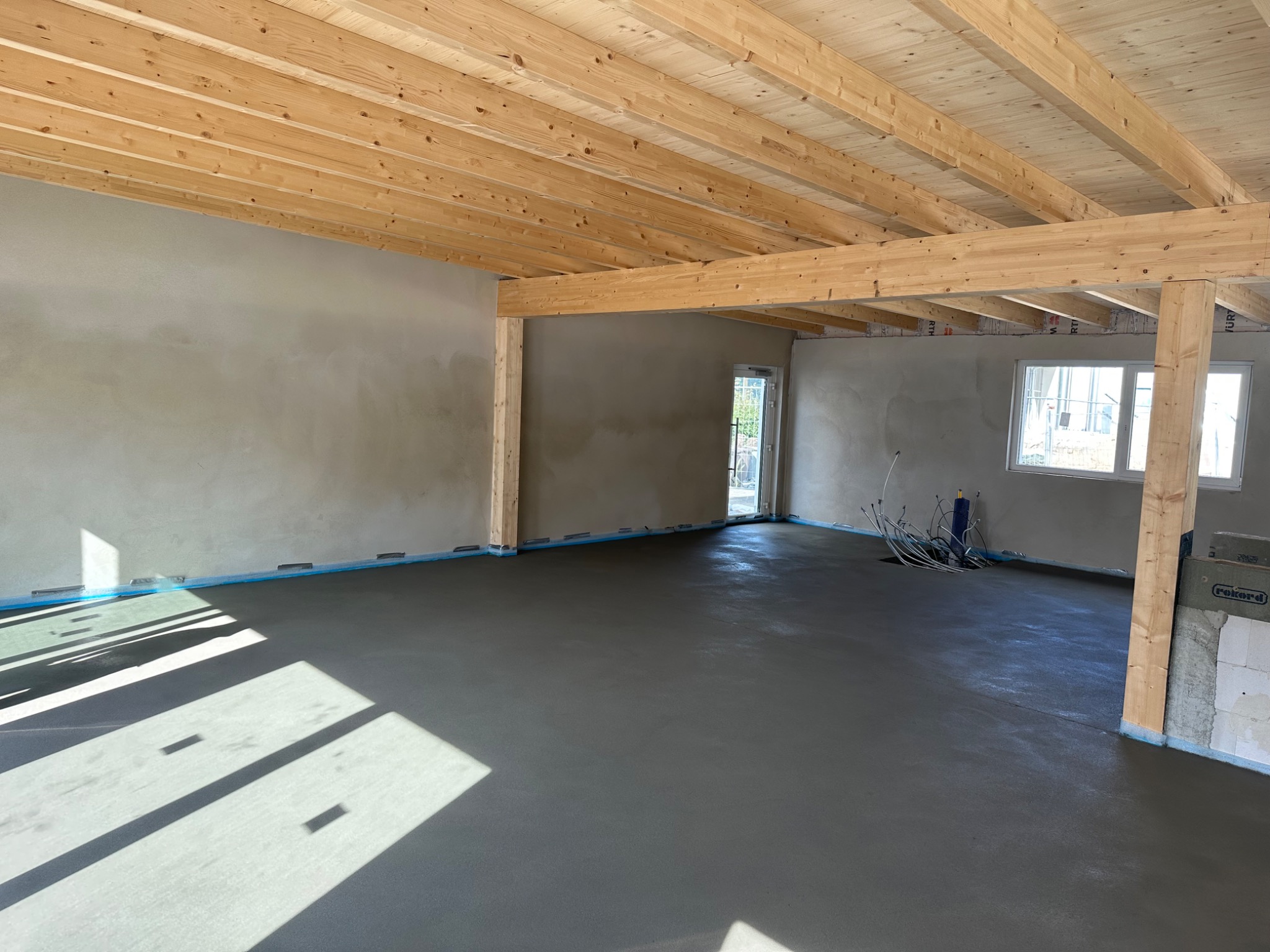 Świeżo wylana, gładka posadzka w surowym wnętrzu z drewnianą konstrukcją stropu i ścianami po wstępnym wykończeniu, widoczne instalacje elektryczne.