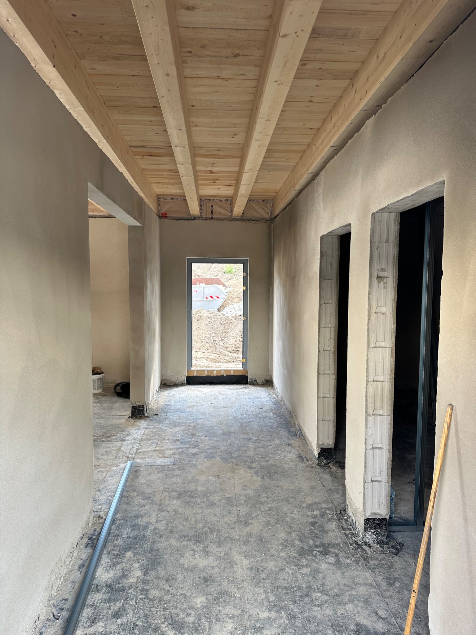 Wnętrze w stanie surowym: świeżo otynkowane ściany, drewniany strop, widoczne otwory drzwiowe z bloków betonowych, przygotowane do dalszych prac wykończeniowych, na podłodze resztki zaprawy.