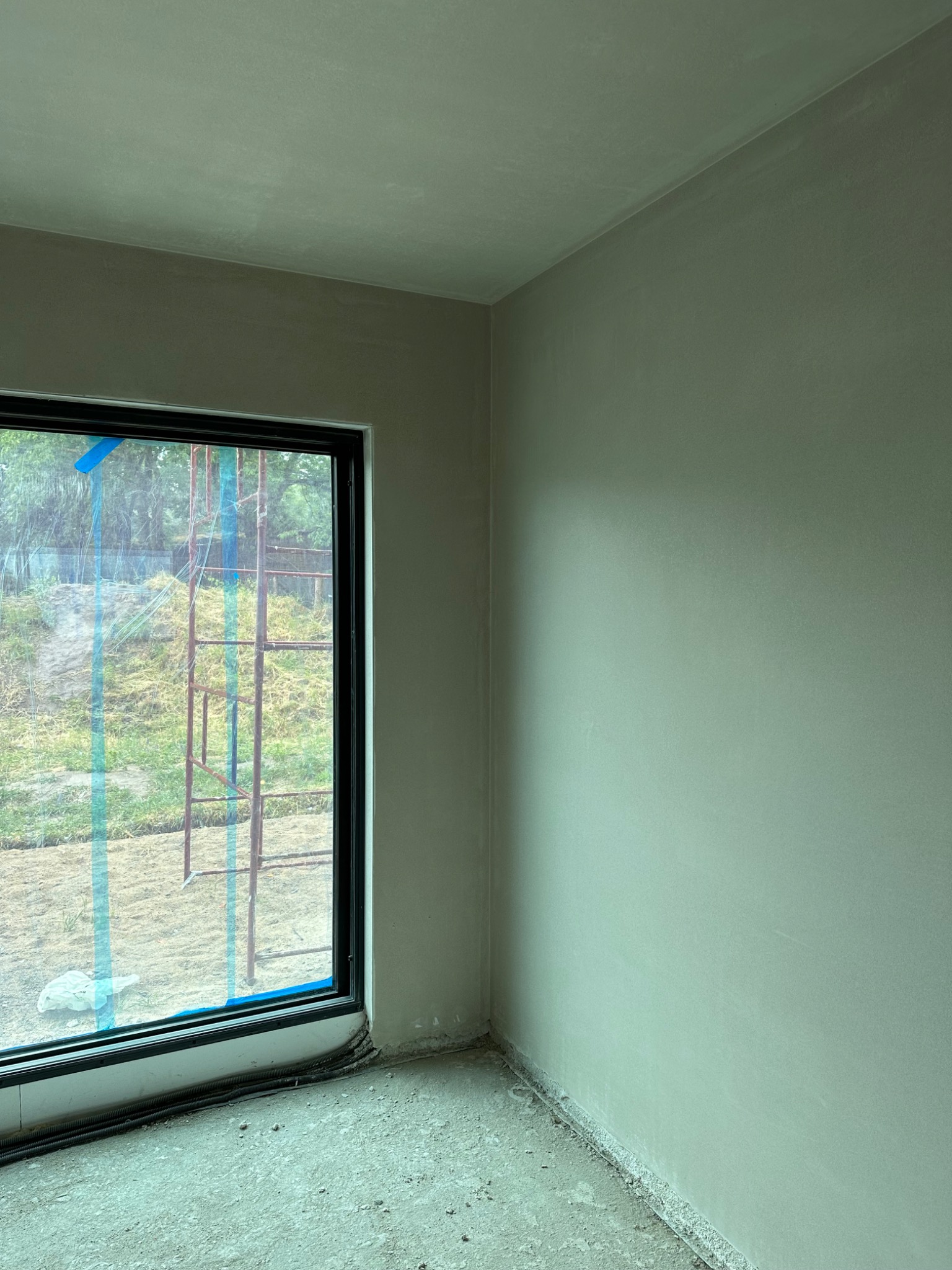 Wnętrze w stanie surowym: świeżo otynkowane ściany w odcieniach szarości, widoczne okno z zabezpieczeniem folią i rusztowaniem na zewnątrz, surowy beton na podłodze.