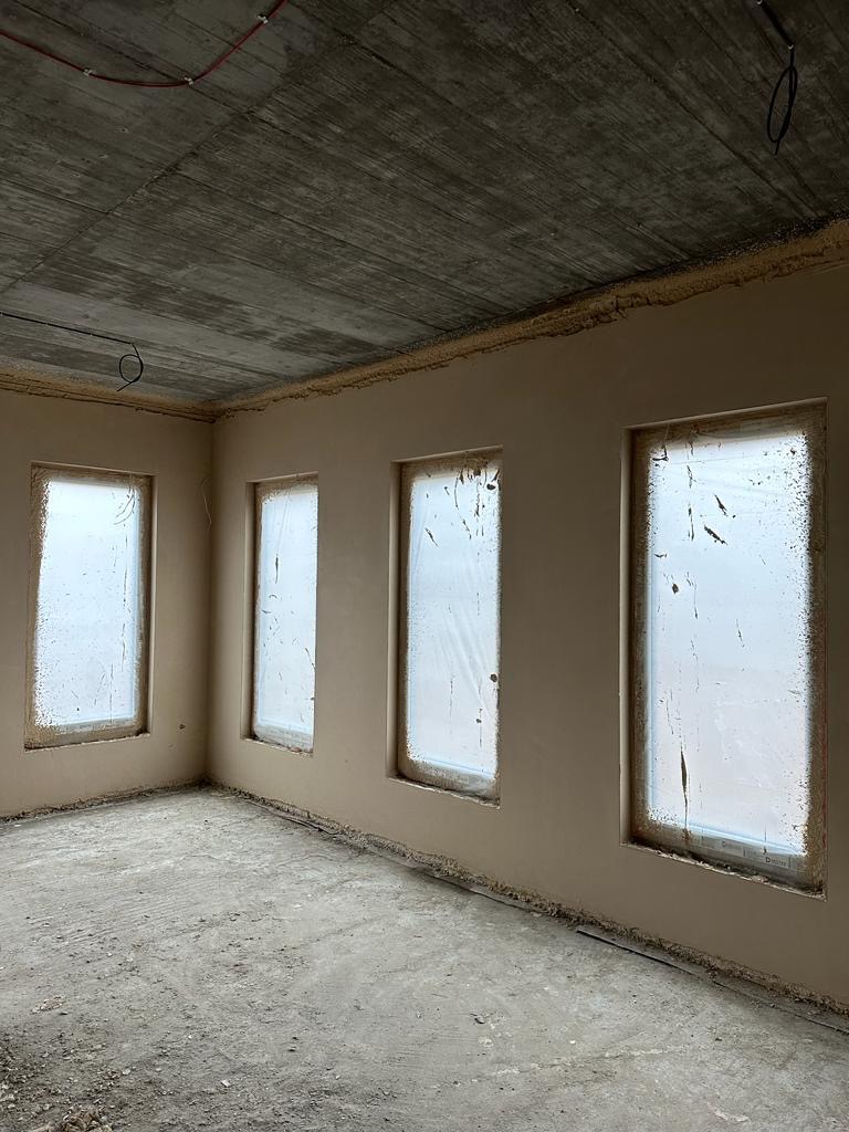 Surowy betonowy sufit w nowym pomieszczeniu z trzema oknami zabezpieczonymi folią, ściany pokryte świeżym, jasnym tynkiem, podłoga betonowa, widoczne przewody na suficie.