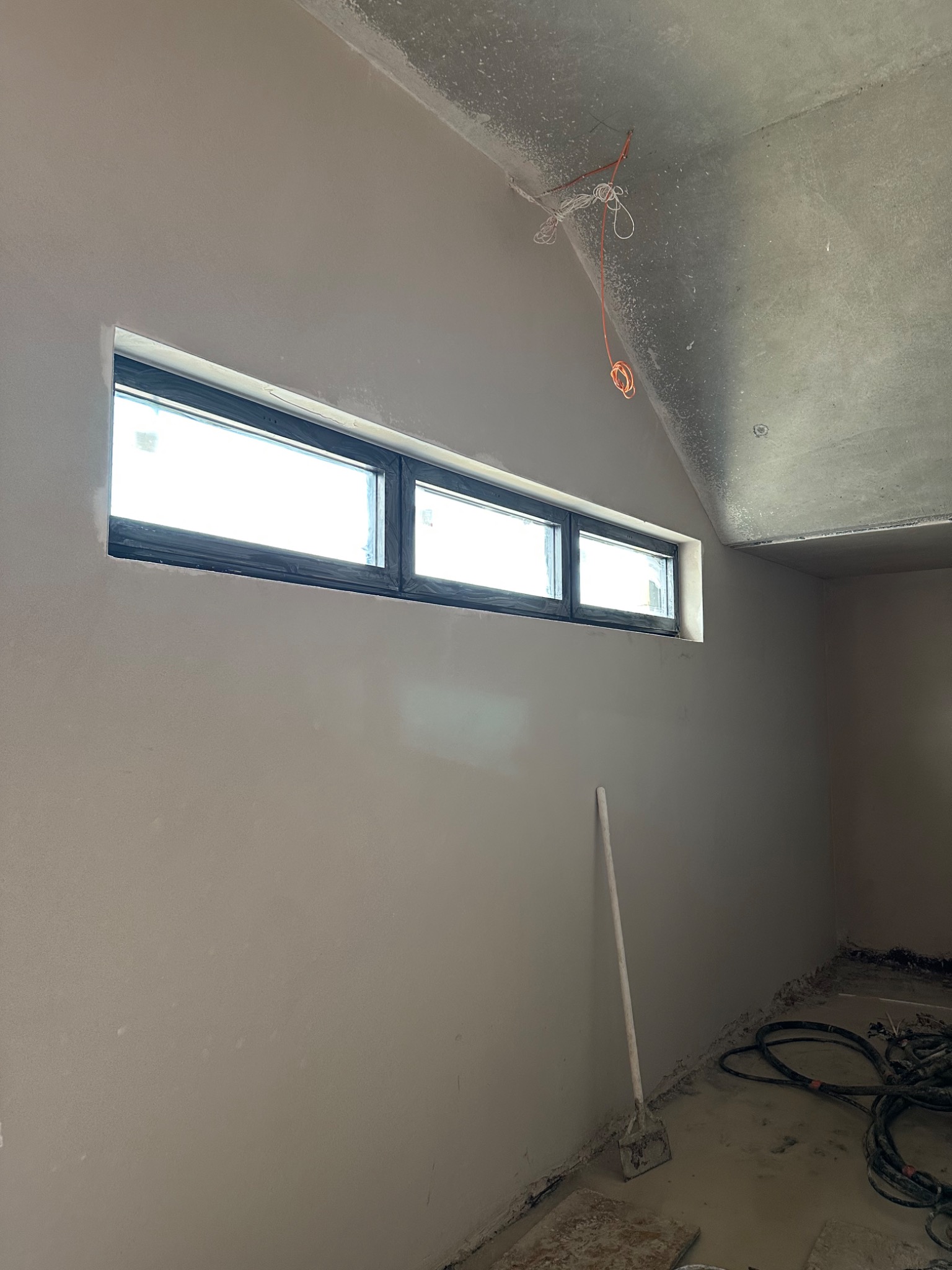 Wnętrze po nałożeniu gładzi, widoczne okno z czarną ramą, przewody elektryczne zwisające z sufitu i narzędzie oparty o ścianę, na podłodze zwinięte kable.