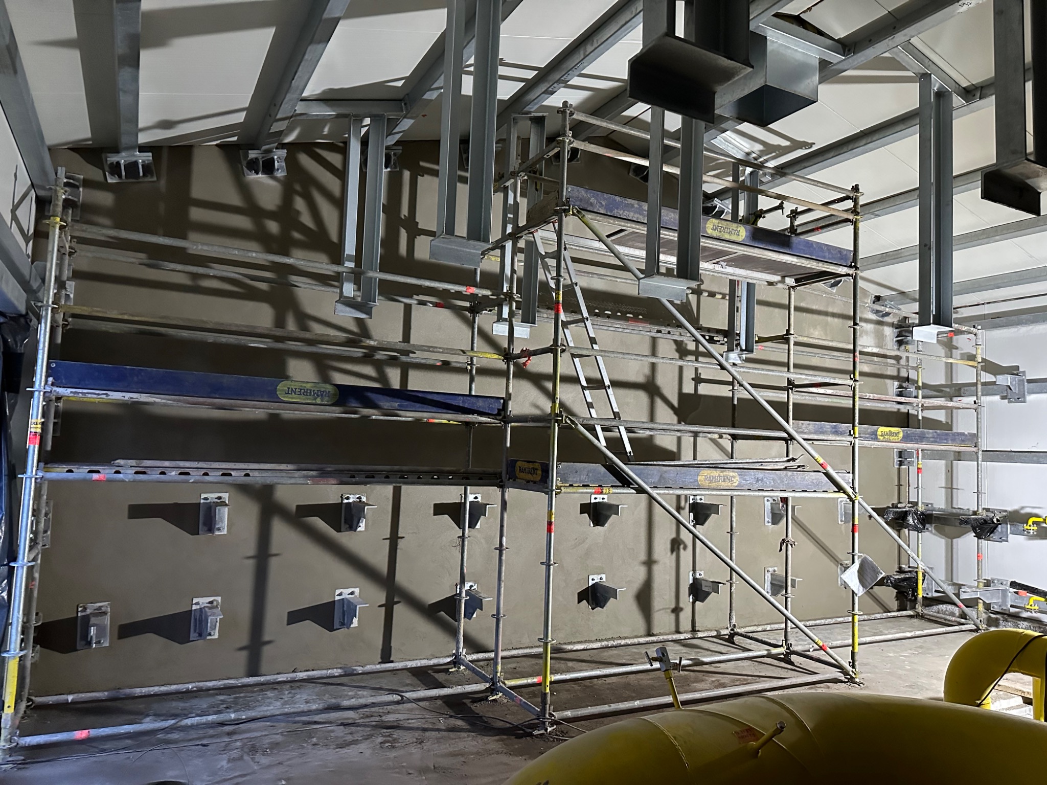 Świeżo nałożony tynk na ścianie wewnątrz budynku, widoczne metalowe uchwyty montażowe i rusztowanie z elementami Ramirent.