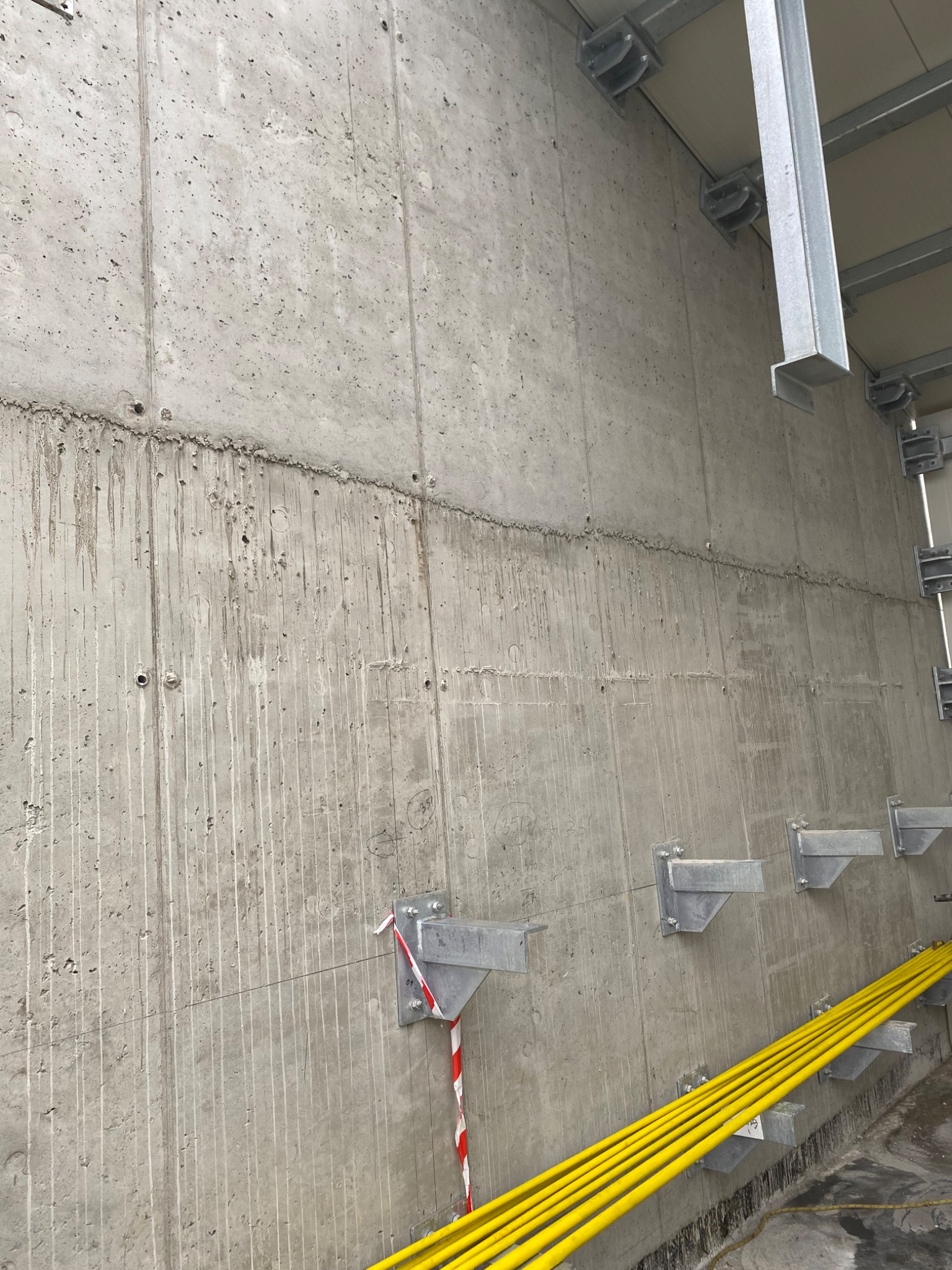 Surowa betonowa ściana z widocznymi śladami po szalunku i zaciekami, metalowe wsporniki przymocowane do ściany, wiązka żółtych kabli ułożona wzdłuż ściany.