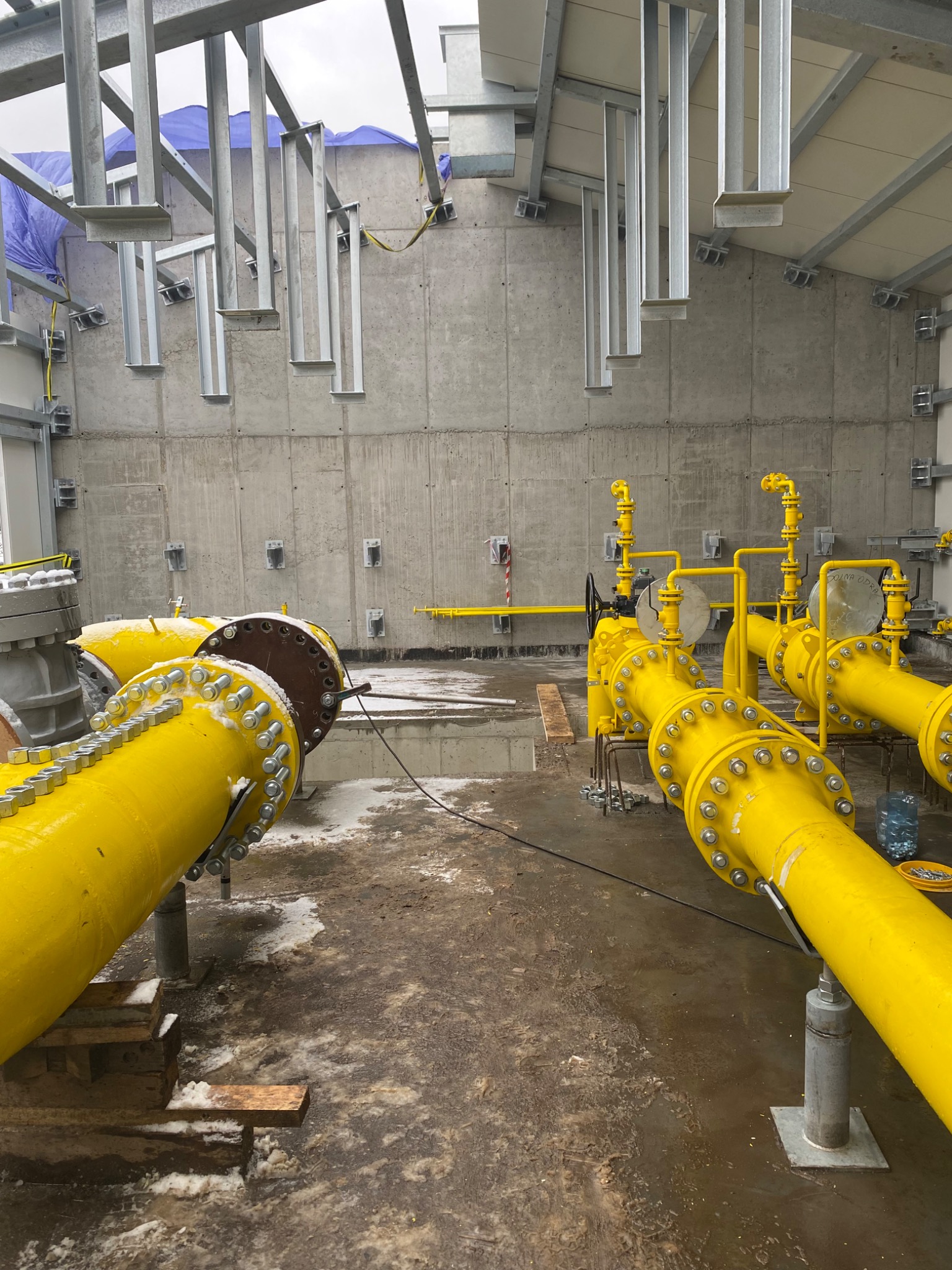 Wnętrze hali z widocznymi, żółtymi rurami gazowymi, wspartymi na metalowych podporach, z betonową ścianą w tle i elementami konstrukcji stalowej pod sufitem.