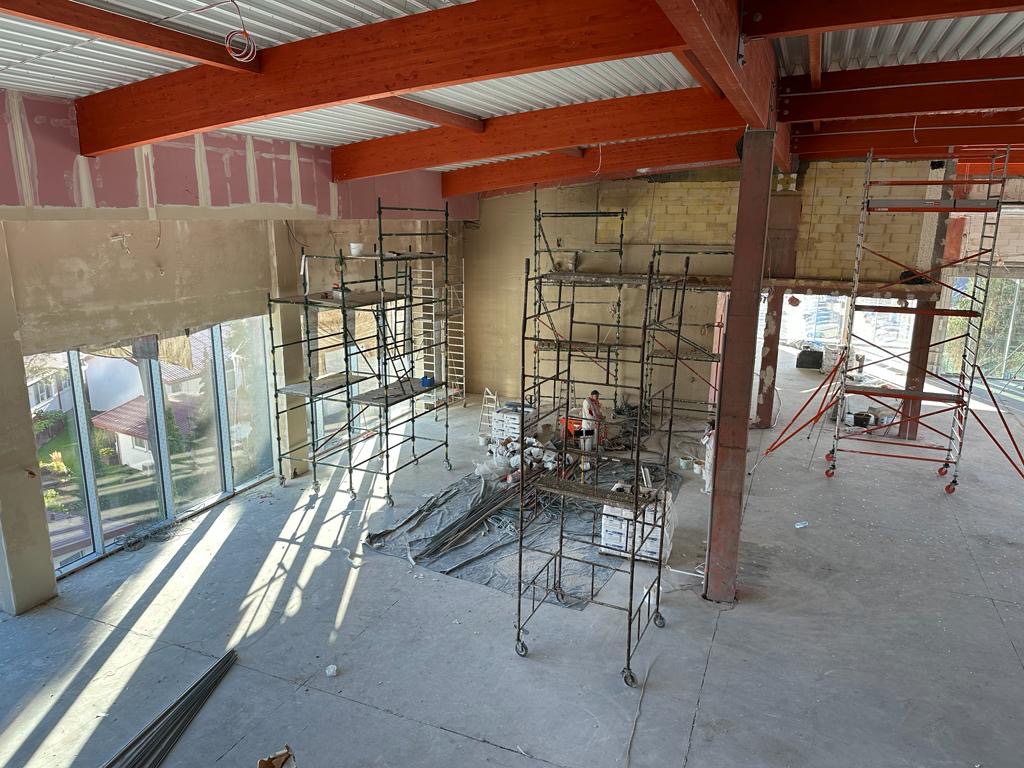 Wnętrze w trakcie remontu: widok z góry na liczne rusztowania, przygotowane ściany pod tynk i duże okna z widokiem na zieleń.