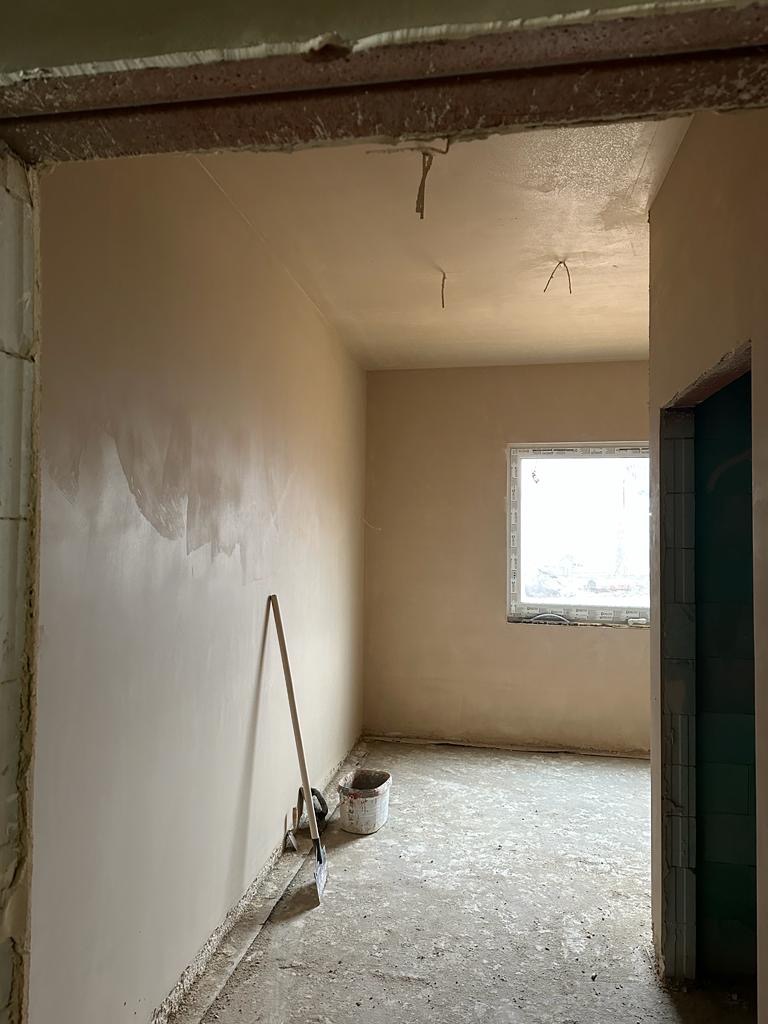 Wnętrze w trakcie prac wykończeniowych: ściany pokryte świeżo nałożoną, jasną warstwą tynku, widoczne narzędzia (szpachelka, wiadro) oparte o ścianę, surowy betonowy strop i podłoga, okno w tle.
