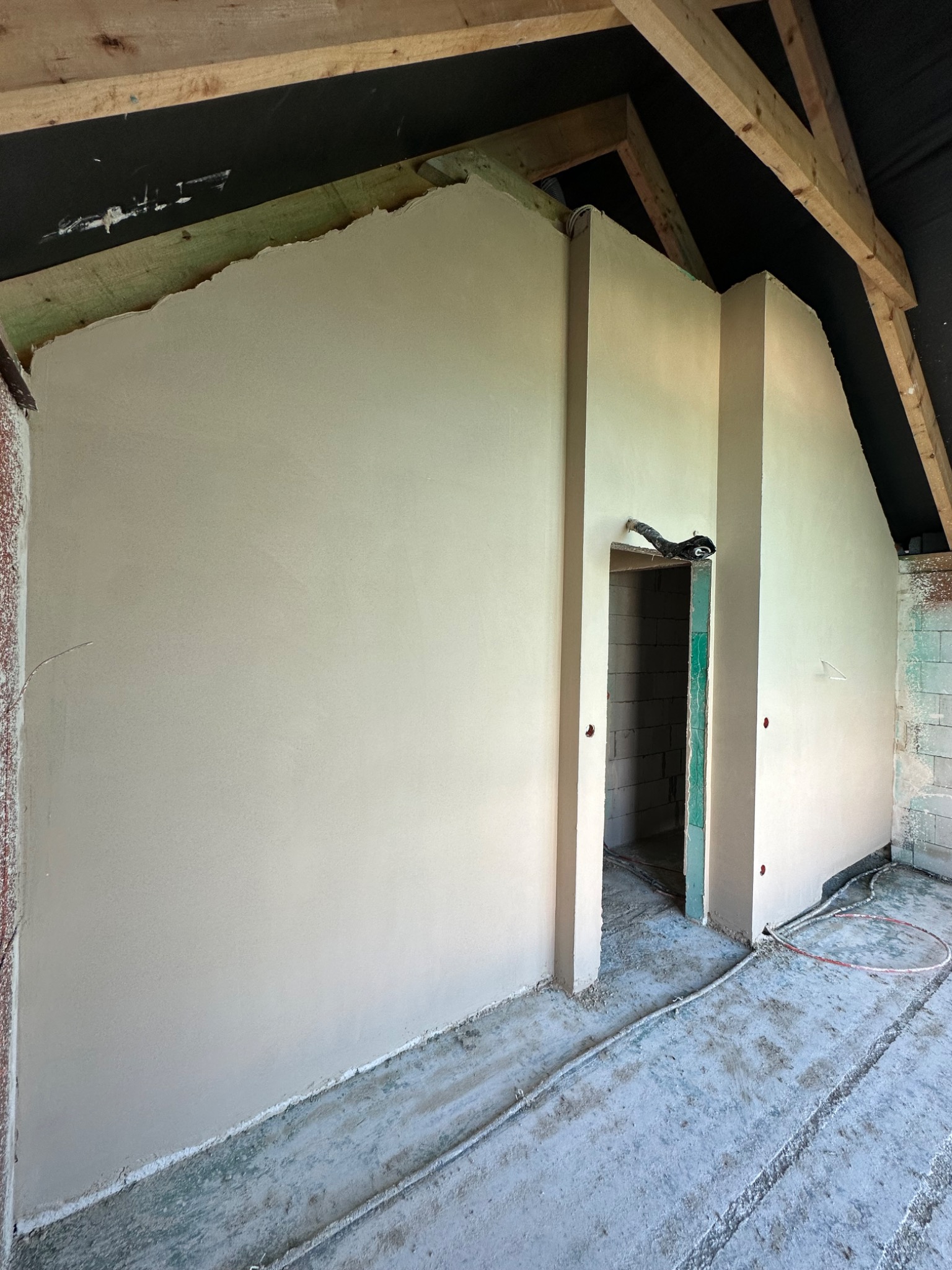 Świeżo otynkowane ściany wewnątrz poddasza, widoczne drewniane elementy konstrukcyjne dachu i instalacja elektryczna na podłodze.