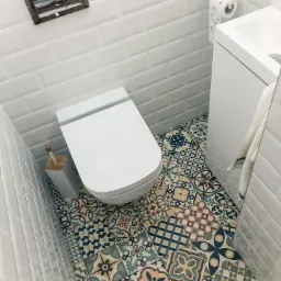 Wysoki kąt widzenia na białą toaletę i umywalkę w małej łazience z białą cegłą na ścianach i dekoracyjnymi płytkami podłogowymi w stylu patchwork.