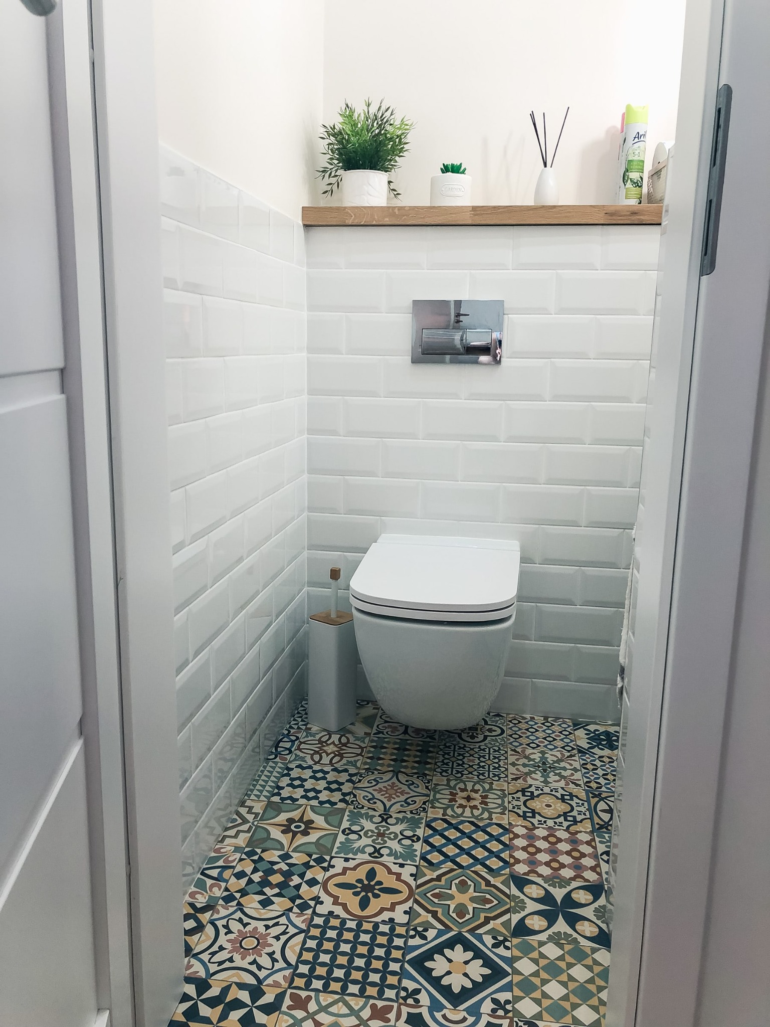 Kompaktowa łazienka z wiszącą toaletą, białe kafelki typu metro, wzorzysta podłoga w stylu patchwork, drewniana półka z roślinami i dyfuzorem zapachowym.