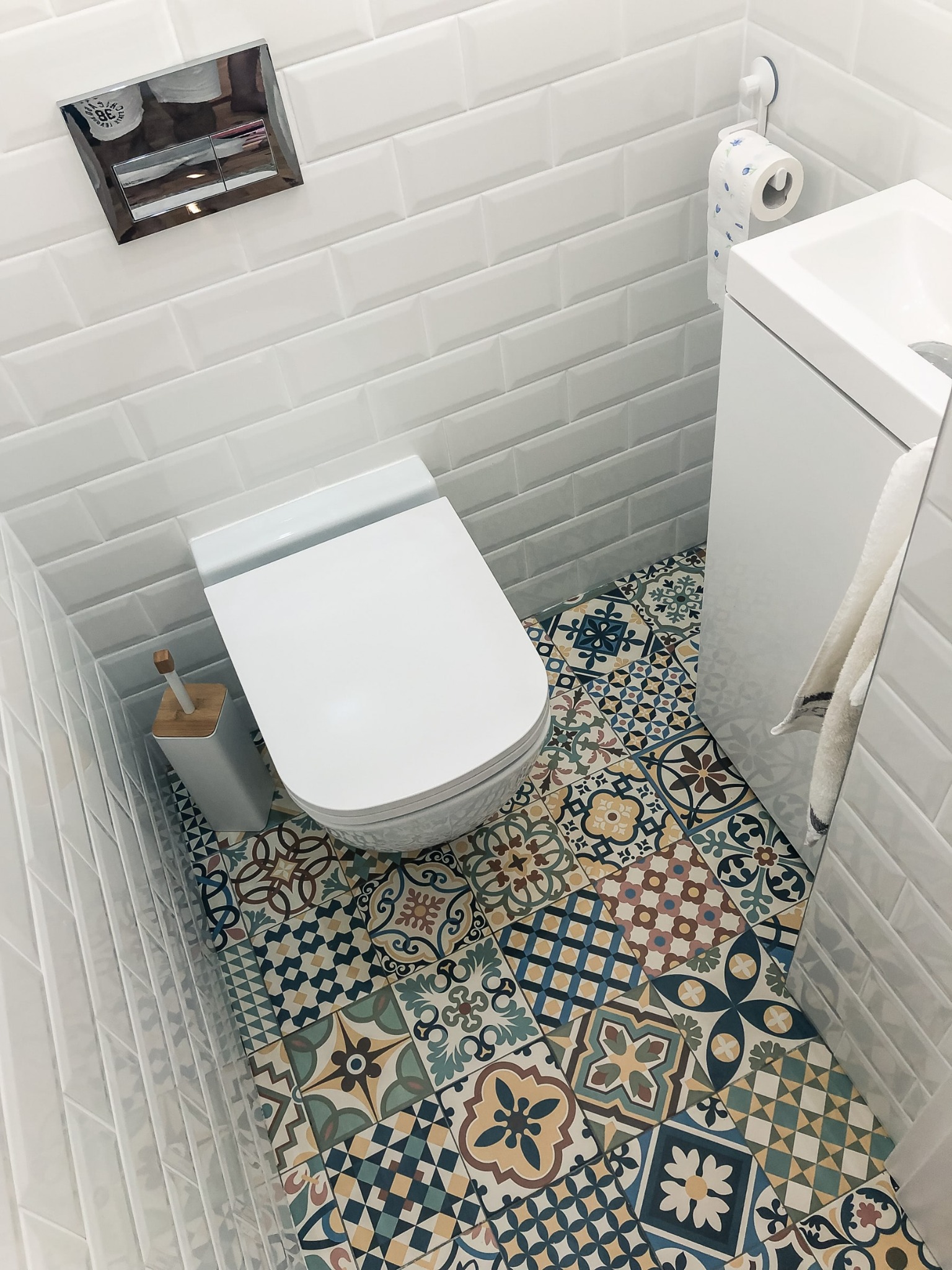 Wysoki kąt widzenia na białą toaletę i umywalkę w małej łazience z białą cegłą na ścianach i dekoracyjnymi płytkami podłogowymi w stylu patchwork.