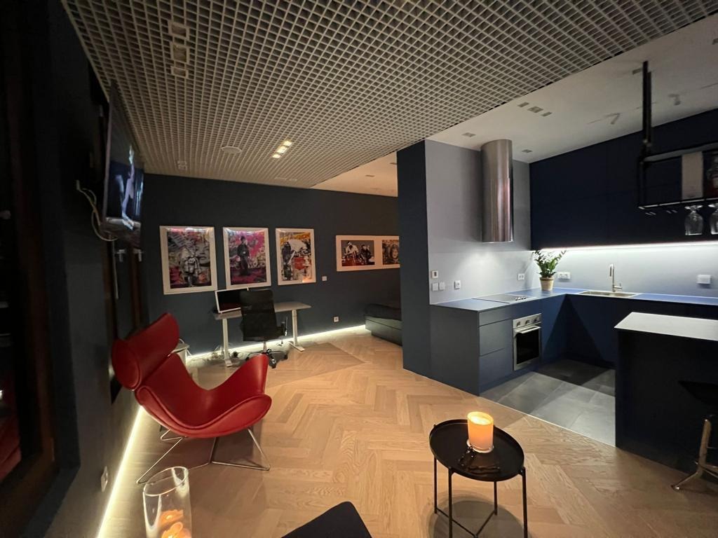 Nowoczesne wnętrze mieszkania z ciemnymi ścianami, parkietem w jodełkę, designerskim czerwonym fotelem i aneksem kuchennym z niebieskimi szafkami oraz metalowym okapem.