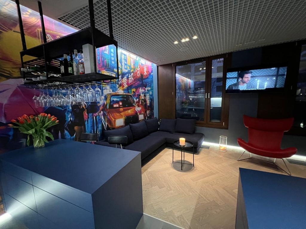 Nowoczesne wnętrze salonu z parkietem w jodełkę, ciemnoniebieską zabudową, sofą narożną, czerwonym fotelem i tapetą z motywem miejskim.