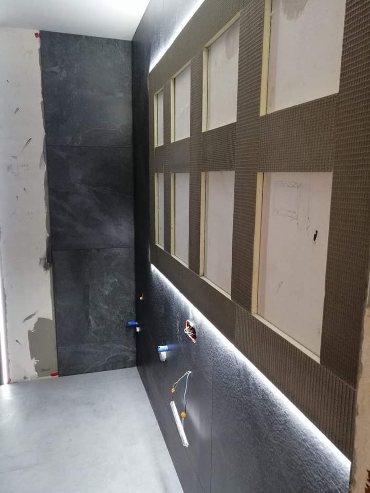 Wnętrze łazienki w trakcie prac wykończeniowych: częściowo ułożone ciemne płytki imitujące kamień, podświetlenie LED wzdłuż ściany, fragment ściany przygotowany pod mozaikę z widoczną siatką...