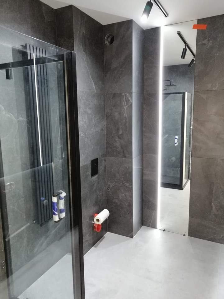 Nowoczesna łazienka z szarymi płytkami imitującymi kamień, kabiną prysznicową z czarnymi ramami, podświetlanym lustrem i minimalistycznym oświetleniem punktowym.