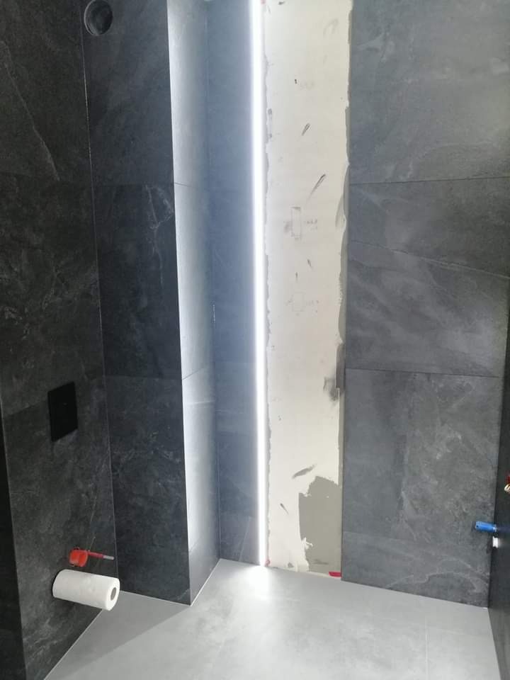 Wnętrze łazienki w trakcie prac wykończeniowych, widoczne szare płytki imitujące kamień, pionowy pas podświetlenia LED oraz fragment ściany przygotowanej do położenia płytek.