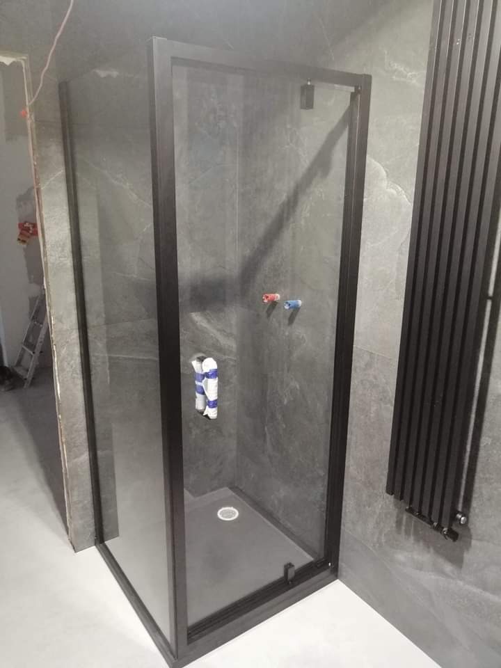 Nowoczesna kabina prysznicowa z czarną ramą i szarymi płytkami imitującymi kamień, widoczne rury zasilające i grzejnik drabinkowy, w trakcie prac wykończeniowych.