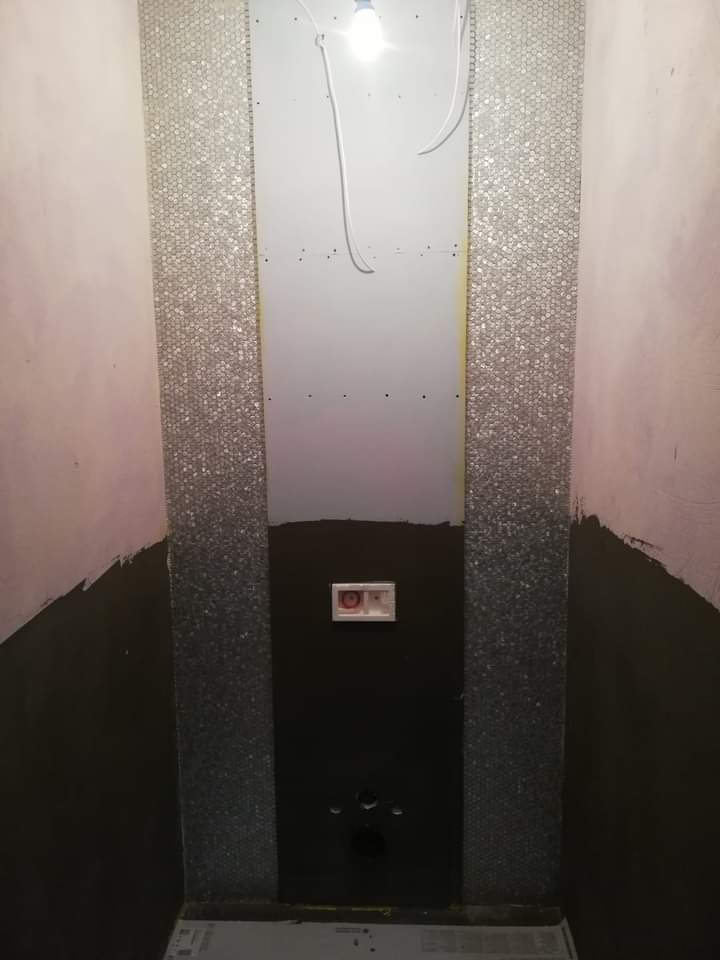 Wnętrze łazienki w trakcie prac: częściowo ułożona mozaika na ścianie, czarna masa uszczelniająca na podłodze i dolnej części ściany, widoczne instalacje wodno-kanalizacyjne.