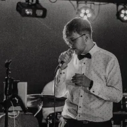 Czarno-białe zdjęcie wokalisty w muszce, śpiewającego do mikrofonu na scenie. W tle widoczne oświetlenie sceniczne i perkusja. Ujęcie z bliska, w półprofilu.