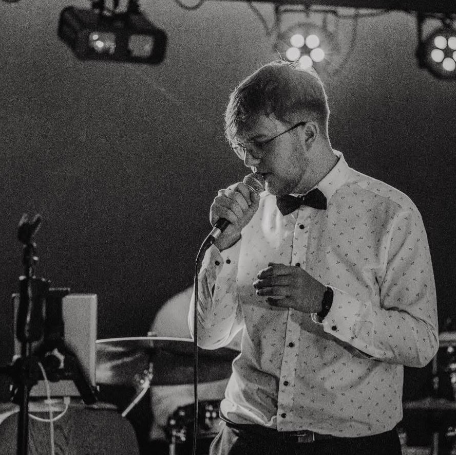 Czarno-białe zdjęcie wokalisty w muszce, śpiewającego do mikrofonu na scenie. W tle widoczne oświetlenie sceniczne i perkusja. Ujęcie z bliska, w półprofilu.