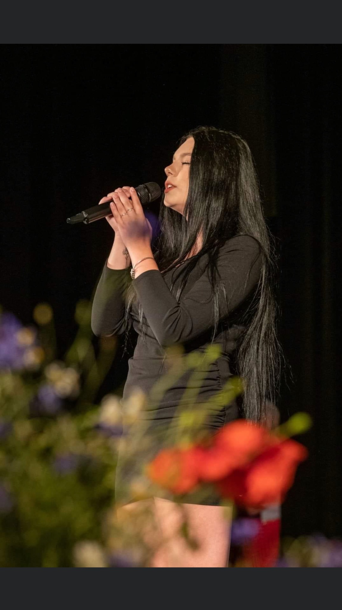 Piosenkarka z długimi, czarnymi włosami śpiewa do mikrofonu, ubrana w czarną sukienkę na tle kwiatów i ciemnej sceny. Widoczny profil twarzy i dłonie z pierścionkami.