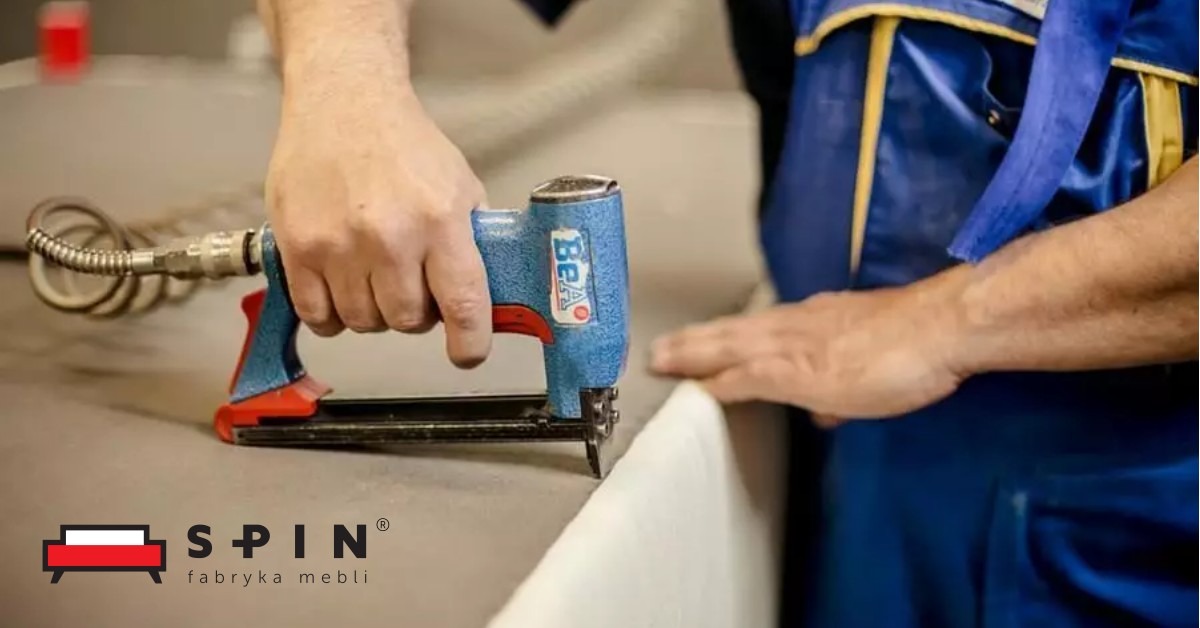 Tapicer używa pneumatycznego zszywacza BEAO do mocowania tkaniny do ramy mebla, widoczne logo producenta mebli w lewym dolnym rogu.