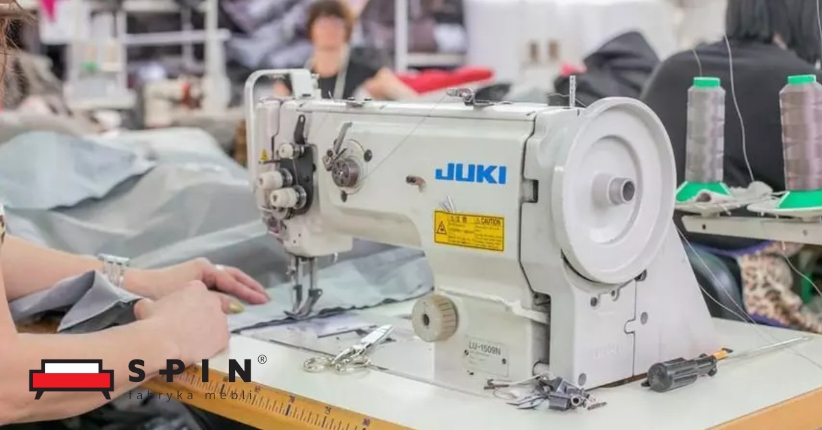 Szwalnia: kobieta szyje materiał na maszynie JUKI LU-1509N, obok leżą nożyczki i miarka, w tle widoczne szpule z nićmi i inne stanowiska pracy.