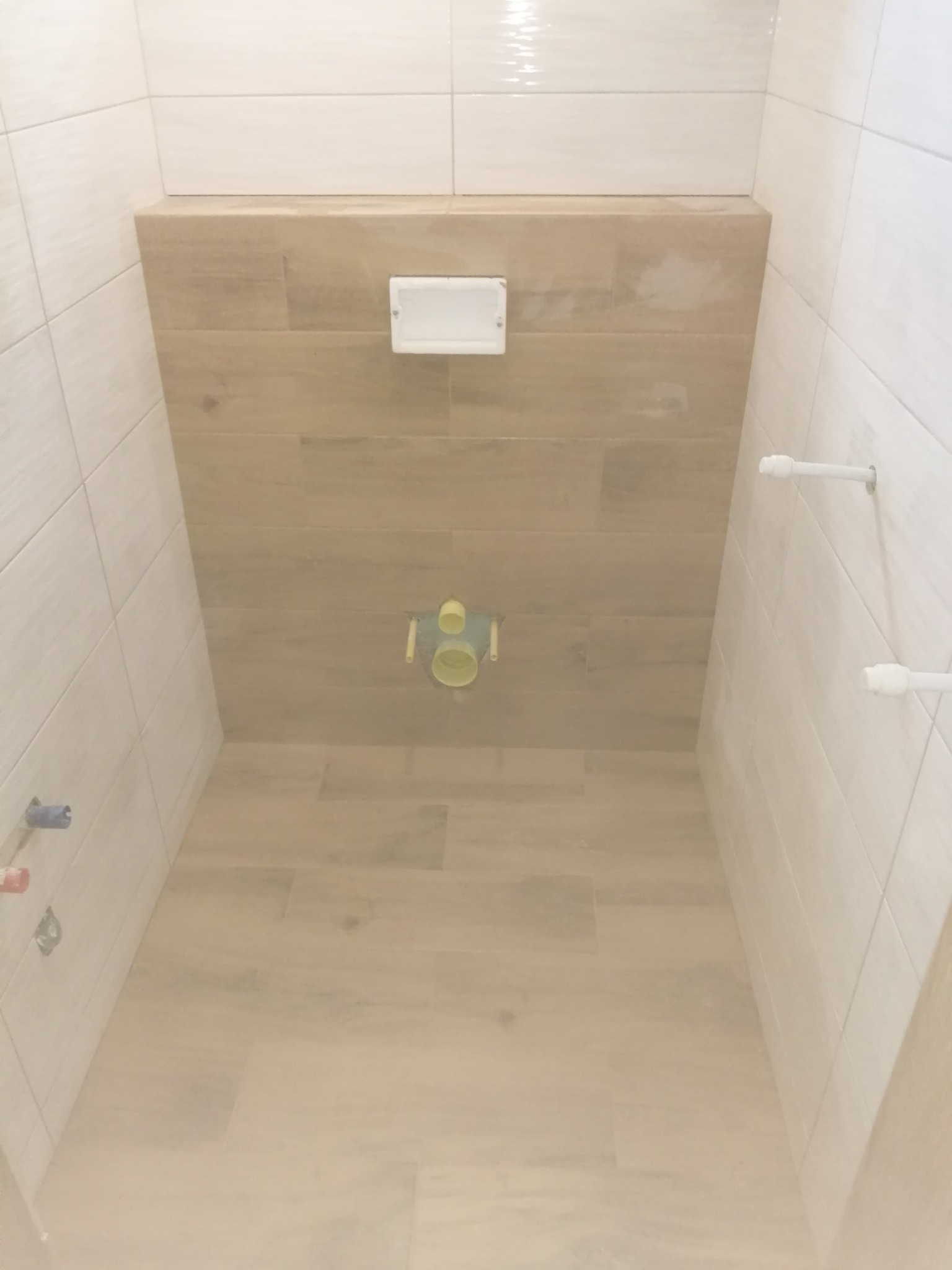 Wnętrze łazienki w trakcie remontu, widoczne białe płytki na ścianach, drewnopodobne płytki na podłodze i zabudowie, oraz elementy instalacji sanitarnej przed montażem ceramiki.