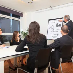 Prelegent rysuje schemat organizacyjny na flipcharcie podczas spotkania biznesowego; uczestnicy siedzą przy stole w jasnym pomieszczeniu.