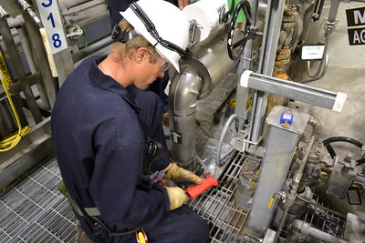 Pracownik w kombinezonie i kasku BHP, kontroluje instalację przemysłową za pomocą miernika, w tle rury i zawory.