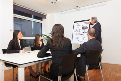 Prelegent rysuje schemat organizacyjny na flipcharcie podczas spotkania biznesowego; uczestnicy siedzą przy stole w jasnym pomieszczeniu.