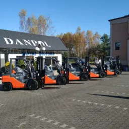 Rząd pomarańczowo-szarych wózków widłowych Toyota na kostce brukowej przed budynkiem firmy Danpol z błękitnym niebem w tle.