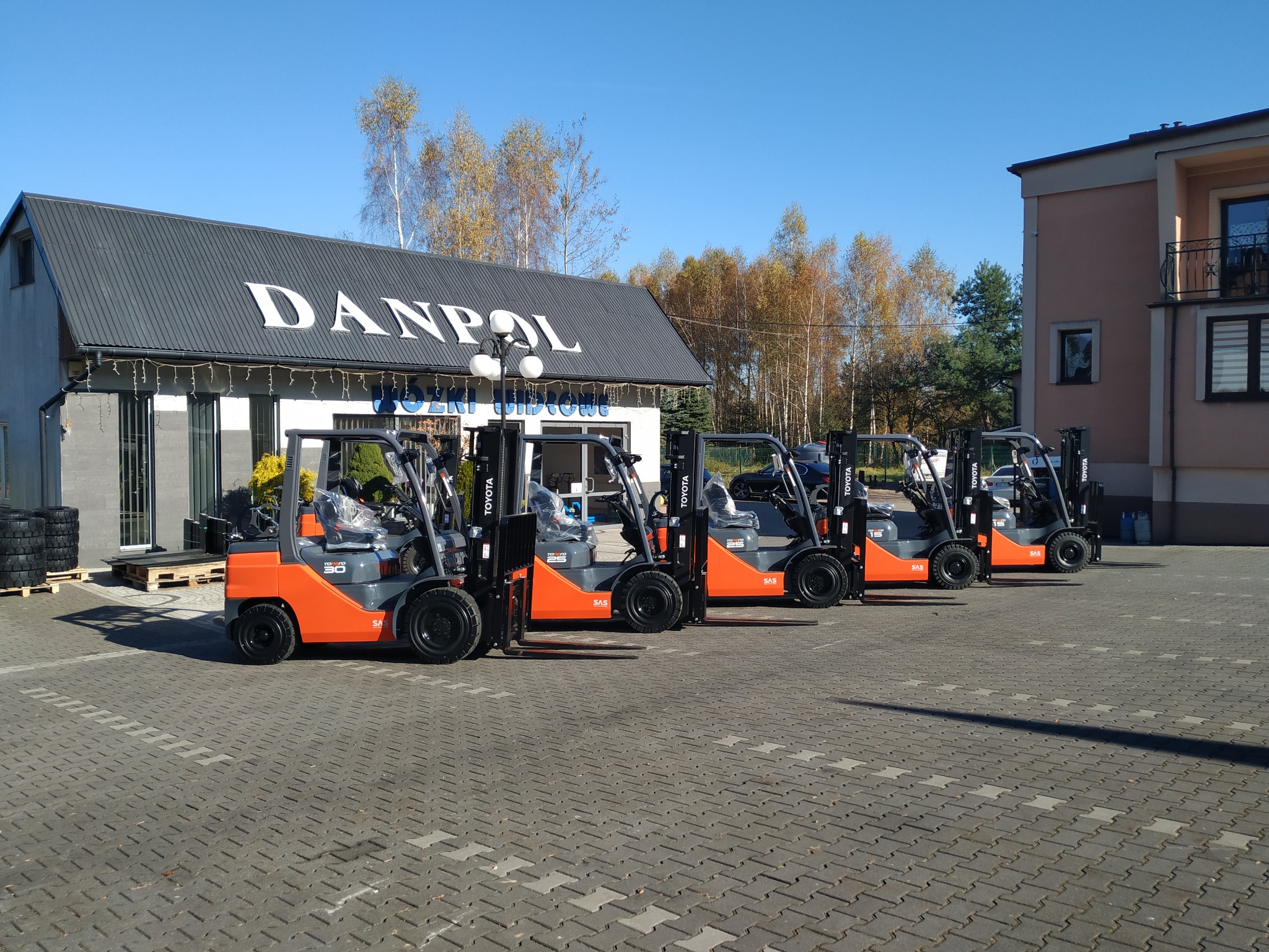 Rząd nowych, pomarańczowo-szarych wózków widłowych Toyota na placu przed budynkiem firmy DANPOL, z oponami ułożonymi na palecie w tle.