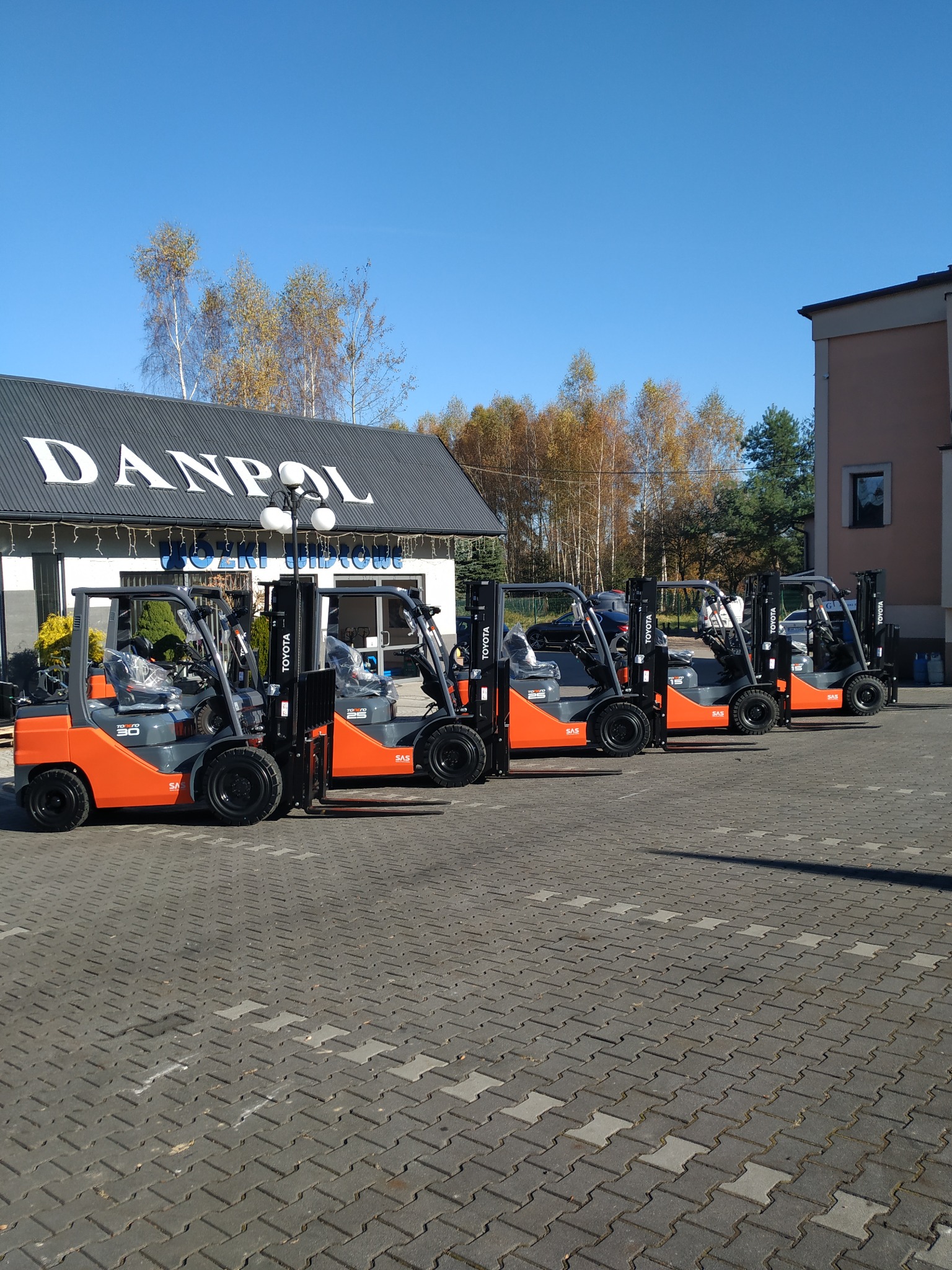 Rząd pomarańczowo-szarych wózków widłowych Toyota na kostce brukowej przed budynkiem firmy Danpol z błękitnym niebem w tle.