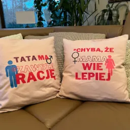 Dwa beżowe dekoracyjne poduszki z nadrukami: po lewej 'Tata ma zawsze rację' z symbolem mężczyzny, po prawej '*Chyba, że Mama wie lepiej' z symbolem kobiety, leżące na brązowej skórzanej kanapie...