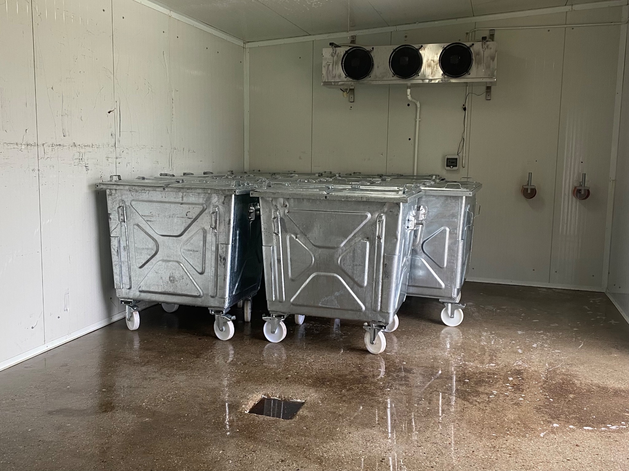 Wnętrze chłodni z trzema metalowymi kontenerami na kółkach, mokra posadzka z odpływem w podłodze, widoczny agregat chłodniczy na ścianie.