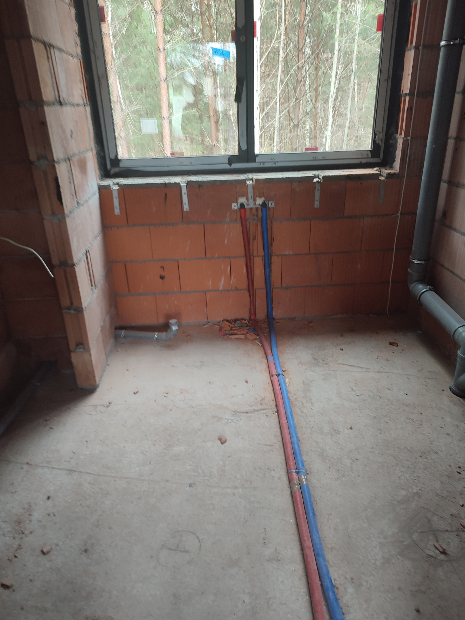 Surowy stan pomieszczenia z widocznymi rurami instalacji wodnej (czerwona i niebieska) wyprowadzonymi ze ściany z czerwonej cegły, przygotowane do podłączenia pod oknem. Widoczne mocowania rur...